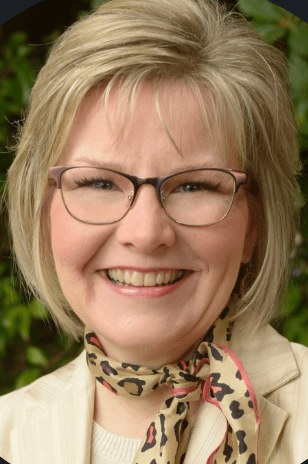 Erica, a smiling woman with a blonde bob, glasses and a patterned neck scarf, photographed outdoors against greenery