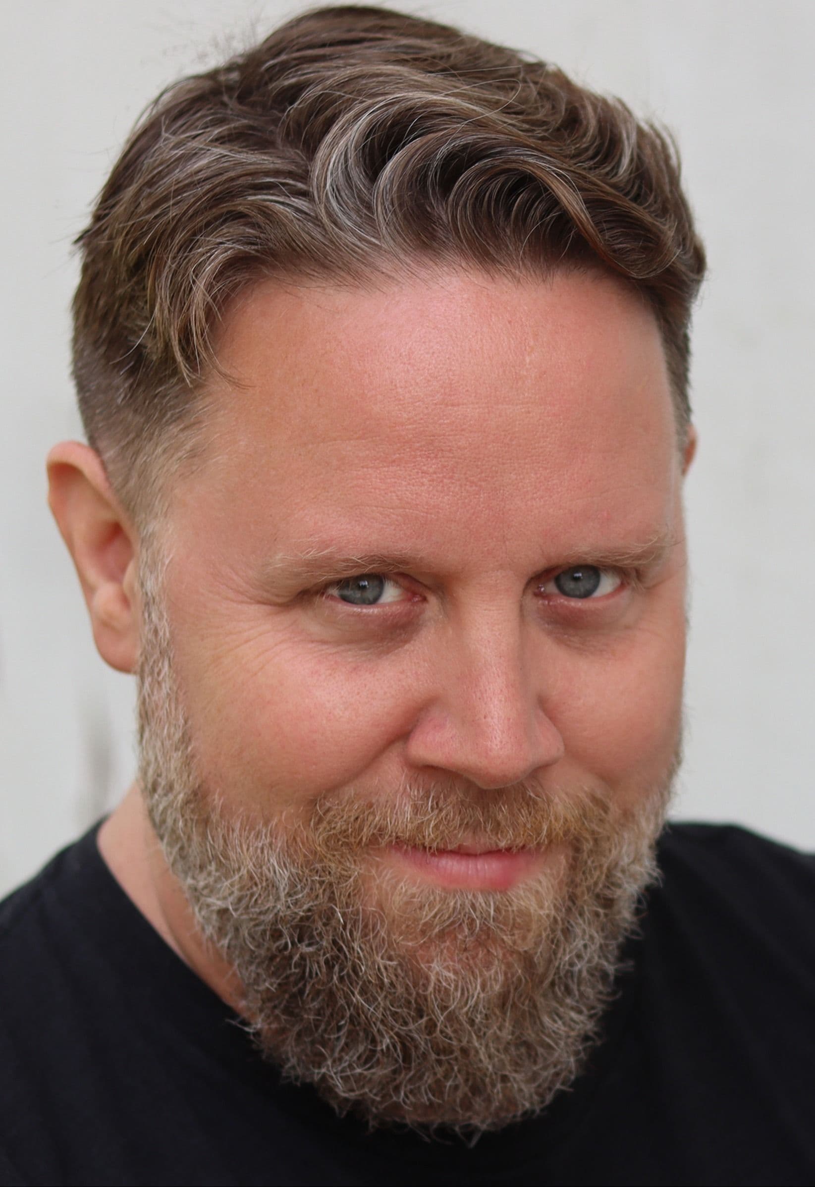 Ken, a bearded man in a black shirt, photographed against a plain neutral wall