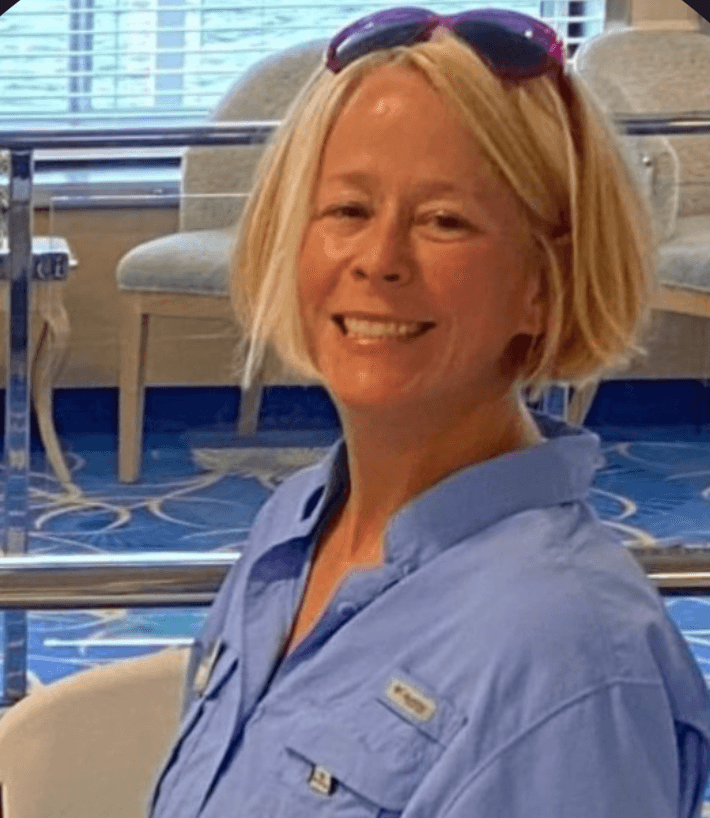Monica, a smiling woman with a blonde bob and sunglasses pushed up on her head, wearing a blue shirt on board a ship